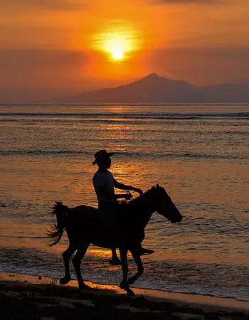 Silhouette of a rider on horseback at sunset on Gili Air, Indonesia, with Mount Agung visible across the Lombok Strait and golden reflections along the shoreline.