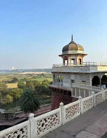 View from Mehtab Bagh in Agra, with a domed Mughal pavilion in the foreground and the Taj Mahal faintly visible across the Yamuna River in the distance.