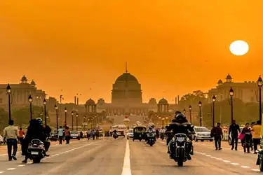 Vibrant sunset view of Rashtrapati Bhavan and Rajpath in New Delhi, India, with people, cars, and motorcycles on the road under an orange sky.