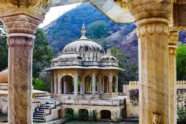 Ornate stone pavilion with a domed roof and carved arches, viewed through two intricately carved columns, set against a backdrop of green hills.