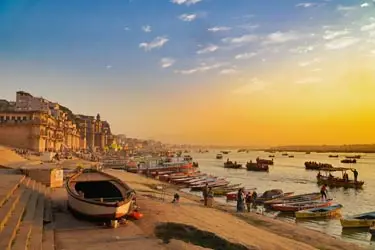 Sunrise over the Ganges River in Varanasi, India, with boats along the ghats, historic riverside buildings, and people gathered along the steps.