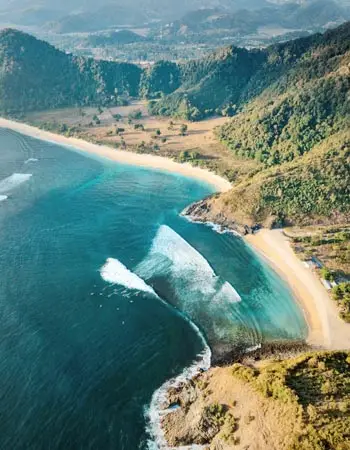 Aerial view of a secluded crescent beach in Lombok, Indonesia, featuring turquoise waves, rolling green hills, and unspoiled tropical coastline.