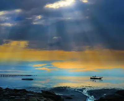 Dramatic sunrise over the sea in George Town, Penang, Malaysia, with sun rays piercing through clouds, a wooden fishing boat floating on calm waters, and a long pier extending into the distance.