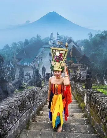 Balinese woman in traditional ceremonial attire ascending temple steps in Bali, Indonesia, with Mount Agung rising in the misty background.