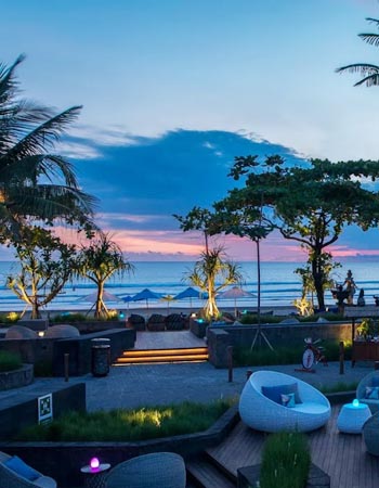 Beachfront lounge at Seminyak Beach in Bali at sunset, with palm trees, soft lighting, and seating areas overlooking the ocean and distant horizon.