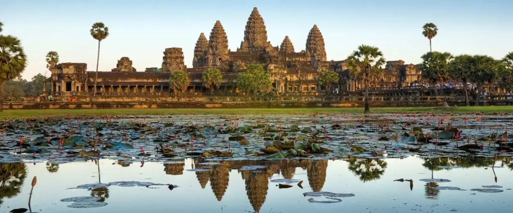 Angkor Wat temple complex at sunrise in Siem Reap, Cambodia, reflected in a lotus-filled pond with palm trees and ancient Khmer architecture.