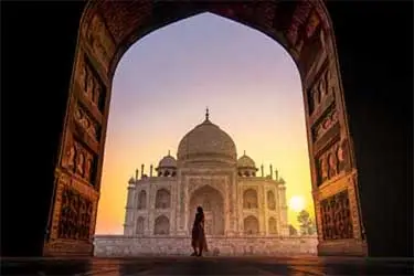 Silhouette of a person framed by an ornate archway, looking toward the Taj Mahal at sunrise with warm golden light illuminating the marble monument.