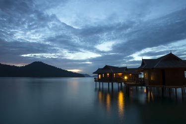 Overwater bungalows at dusk along a tranquil island coastline in Malaysia, reflecting softly on calm blue waters under a dramatic evening sky