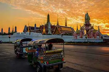 Tuk-tuk driving past the Grand Palace in Bangkok, Thailand at sunset, with golden temple spires glowing against an orange evening sky.