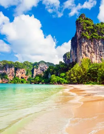 White-sand beach in Krabi, Thailand, with turquoise Andaman Sea waters and towering limestone cliffs rising above lush tropical greenery.