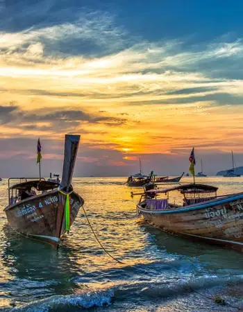 Long-tail boats anchored off the coast of Krabi, Thailand at sunset, with golden light reflecting across calm Andaman Sea waters and a colorful evening sky.