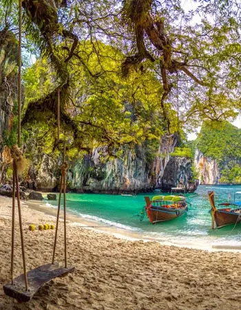 Secluded beach in Krabi, Thailand with a wooden swing beneath tropical trees, long-tail boats anchored offshore, and dramatic limestone cliffs framing turquoise waters.