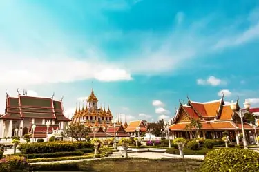 Grand Palace complex in Bangkok, Thailand, featuring traditional Thai temple buildings, ornate rooftops, and manicured gardens under a clear blue sky.