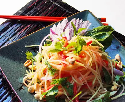 Thai papaya salad (som tam) served on a black plate, featuring shredded green papaya, chili, peanuts, lime, and fresh herbs in a vibrant traditional dish.