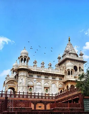 Ornate marble palace in Jodhpur, India, featuring domes, carved balconies, and intricate architectural detailing, set against a clear blue sky with birds in flight.
