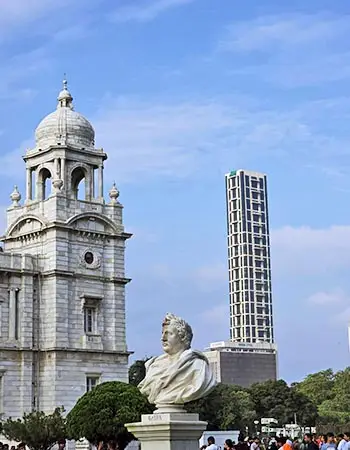 Victoria Memorial in Kolkata, India, with its white marble dome and statue in the foreground, set against a modern high-rise building and blue sky.