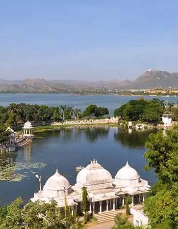 Scenic view of Udaipur with a tranquil lake, domed white pavilions, lush greenery, and surrounding hills under a clear sky.