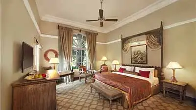 Elegant guest room at Taj Hari Mahal, Jodhpur, featuring a four-poster bed, classic wooden furnishings, warm lighting, and a large arched window with garden views.