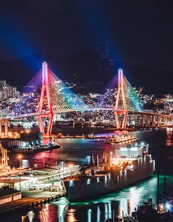 Gwangan Bridge illuminated in vibrant colors over Busan Harbor at night, highlighting the modern skyline and coastal cityscape of Busan, South Korea.