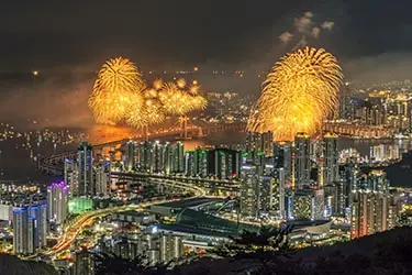 Fireworks illuminating the skyline and coastline of Busan, South Korea during a vibrant festival, with city lights reflecting along the waterfront.
