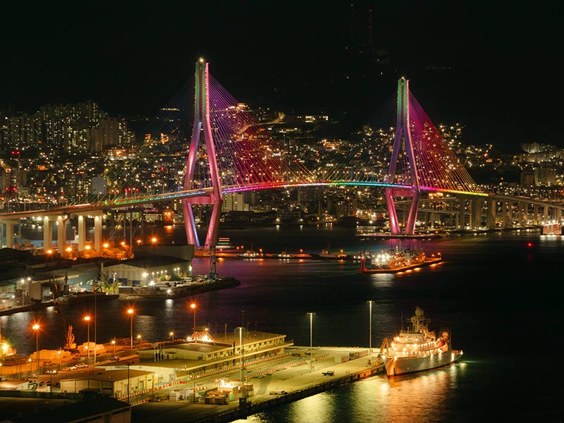 Night view of Busan Harbor Bridge illuminated in colorful lights, spanning over the port with ships, waterfront terminals, and city skyline glowing in South Korea.