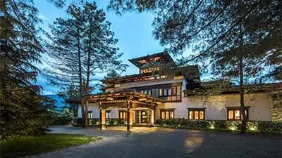 COMO Uma Punakha in Bhutan, featuring a traditional Bhutanese-style lodge surrounded by pine trees, warmly lit at dusk with a serene mountain setting.