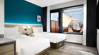 Modern twin guest room at Four Points by Sheraton Josun Seoul, featuring two white beds, teal accent wall, minimalist wood furnishings, and a large window with outdoor terrace seating.
