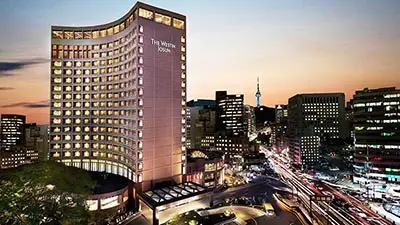 The Westin Josun Seoul hotel exterior at dusk, a curved high-rise illuminated with warm lights overlooking a bustling city street and skyline near Myeongdong, Seoul.