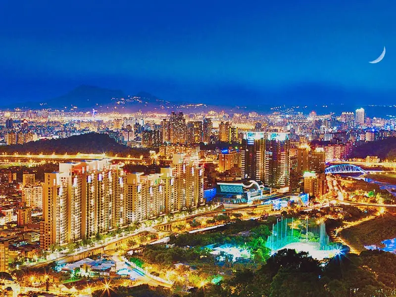 Night view of Taipei city skyline glowing with lights, featuring dense high-rise buildings, illuminated roads, riverside park, and a crescent moon over the urban landscape.