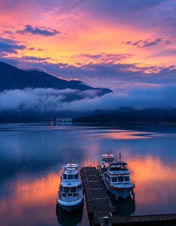 Sun Moon Lake at sunset in Taiwan, featuring tranquil water reflecting vivid pink and purple skies, mist drifting across mountain silhouettes, and small boats docked beside a quiet lakeside pier.