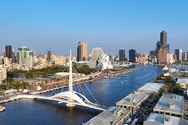 Kaohsiung skyline along the Love River waterfront in Taiwan, featuring modern skyscrapers, harbor views, and a sweeping pedestrian bridge under clear blue skies.
