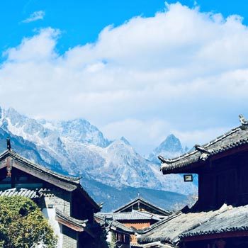 Traditional tiled rooftops in Lijiang Old Town, Yunnan, with the jagged peaks of Jade Dragon Snow Mountain rising dramatically in the background under a clear blue sky.
