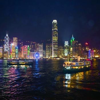Hong Kong skyline at night viewed from Victoria Harbour, with illuminated skyscrapers, colorful city lights, and a ferry crossing the harbor waters.