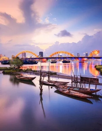 Vietnam_350-450_Danang_Dragon_Bridge Dragon Bridge glowing at dusk over the Han River in Da Nang, Vietnam, with traditional wooden boats in the foreground capturing the city’s blend of modern design and coastal charm.